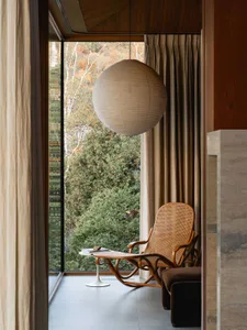 A minimalist residential room featuring a natural wood chair, a spherical pendant light, and a view of greenery outside throu