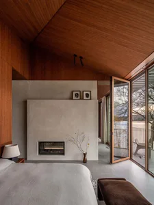 A modern bedroom with a concrete fireplace and large windows overlooking a serene outdoor view.