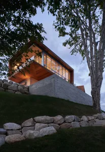 A modern house with a concrete exterior and large windows, surrounded by rocky ground and trees at dusk.