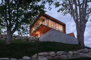 A modern house with wood exterior and concrete foundation, featuring large windows and a sloped roof.