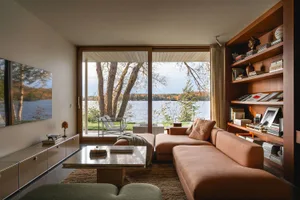 A modern living room featuring natural light from a large glass door overlooking a scenic lake. The room includes a comfortab