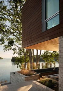 Modern residential exterior featuring wood siding, stone accents, and large glass windows overlooking water and greenery.