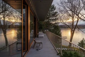 Modern exterior of a waterfront residence with wooden deck and furniture, overlooking a serene lake with sunset reflections.
