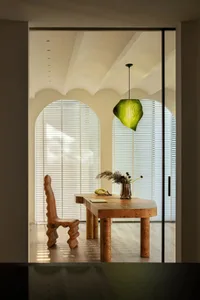 A modern home office featuring a wooden desk and chair, with a green pendant light and large windows.