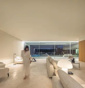 A modern living room featuring concrete walls and a glass wall with a view of a pool area at night.
