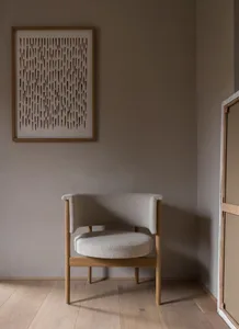 A modern interior fabric chair with wooden legs and arms, placed against a neutral-colored wall with a framed artwork.
