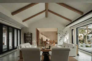 A modern living room with white sofas, wooden beams on the ceiling, and a glass wall overlooking an outdoor area.