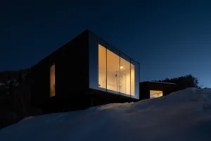A modern metallic hillside house illuminated at night with snow-covered ground and surrounding trees.