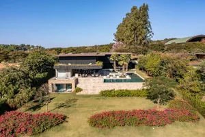 A modern residential home with a stone exterior, glass walls, and a large pool surrounded by greenery.