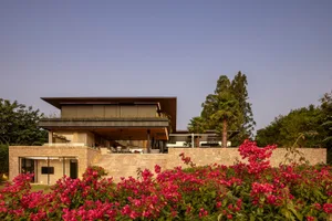 A modern residential house with a sandstone and concrete exterior, surrounded by lush greenery and red flowers.