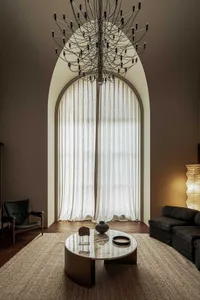 A modern living room featuring neutral-toned, organic materials and a large, arched window with white curtains.