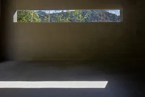 A minimalist interior space with a concrete floor and a small, rectangular window overlooking trees and mountains.