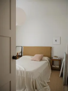A minimalist bedroom with a bed covered in beige linens, a pink pillow, and a simple headboard.