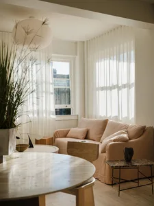 A minimalist living room featuring a beige sofa, round marble tables, and a potted plant by a window with sheer curtains.