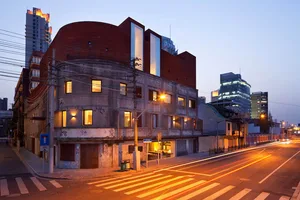 A city street corner at dusk with a modern building featuring concrete facade and illuminated glass windows.