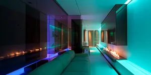A modern living room with glass walls and colorful lighting features a sleek green sofa and a futuristic fireplace.