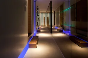 An illuminated, minimalist corridor featuring wooden benches, concrete floor, and glass walls