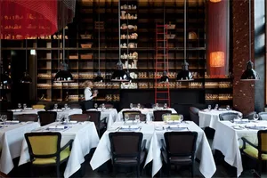 Modern restaurant interior with chairs and tables set with white cloth, multiple pendant lights, and red brick walls.