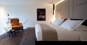A minimalist bedroom featuring a large bed with a white cloth, an orange armchair, and a wooden floor with a gray rug.