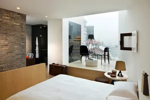 A minimalist bedroom with white furniture, a wood headboard, and a view of the city skyline through a large window.