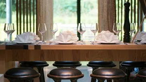 A neatly arranged restaurant table setting with wooden placemats, white napkins, and clear wine glasses, surrounded by black