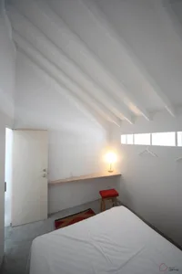 A minimalist bedroom featuring a white bed with a red stool and a lit lamp, under a sloped white ceiling with wooden beams an