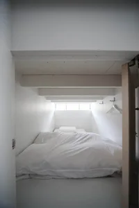 A minimalist bedroom with a bed covered in white textiles and a small window on the ceiling.