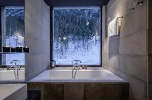 A modern concrete bathroom with a large window overlooking a snowy forest.