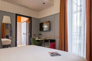A modern hotel room with a white bed, a bedside table with a red magazine and a green chair, and an orange curtain.