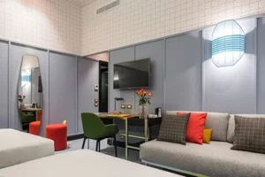 A modern hotel room with a grey upholstered sofa, orange cushions, and a television mounted on the wall.