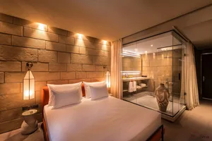 A minimalist bedroom with a cement-textured wall, white bedding, and a glass-enclosed bathroom.