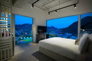 A minimalist bedroom with a large white bed overlooking a cityscape at dusk, featuring sleek, modern furniture and a large wi