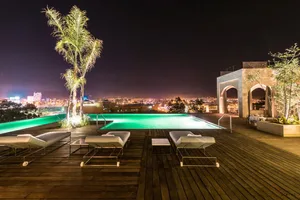 Modern minimalist pool deck with a night view, featuring a wooden deck, white lounge chairs, and palm trees, overlooking a vi
