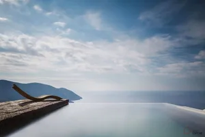 An infinity pool with a metal lounge chair overlooking a scenic landscape with mountains and the ocean.