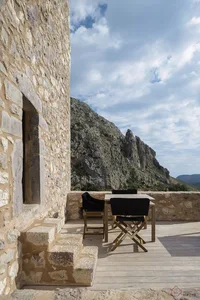 A modern stone and wood residential dwelling with a table and chairs on a deck overlooking rugged mountains.