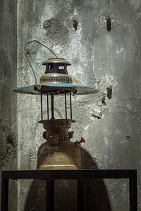 An antique, metal lantern with a glass chimney and handle, placed on a wooden shelf against a worn, concrete wall.