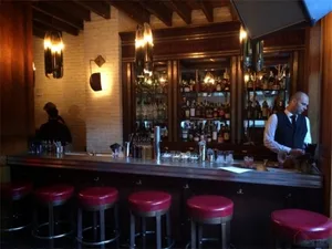 A warm-toned wooden restaurant bar with red stools, illuminated by hanging lights and surrounded by shelves filled with bottl