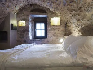 A cozy bedroom with a white bed and stone walls, featuring a small window with natural light.