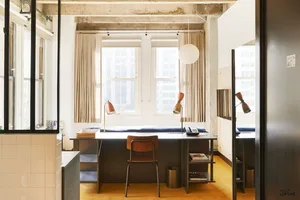 A minimalist office desk space featuring a wooden chair, desk lamp, and computer monitor with a window view of city buildings