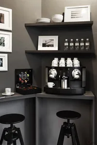 A minimalist coffee bar featuring a coffee machine with white cups and saucers on a black countertop, black stools, and decor