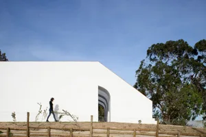 A modern residential building with a white concrete exterior, features large windows and an open archway, set against a clear