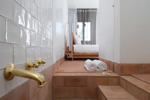 A minimalist bedroom featuring white tiled walls and terracotta tiles on the floor, with a bed and towels.