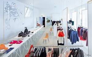 A modern clothing store with white walls, wooden chairs, and clothes displayed on racks.