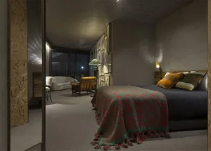 A bedroom with minimalist cement walls, a bed with a red and green blanket, and a small sofa in the background.