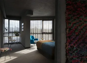 A modern bedroom featuring a concrete wall with embedded hyphae, a blue armchair, and a desk with a plant.