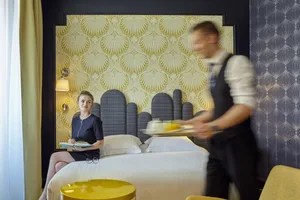 A woman sits on a bed in a hotel room decorated with patterned wallpaper, a leather headboard, and a cotton blanket.