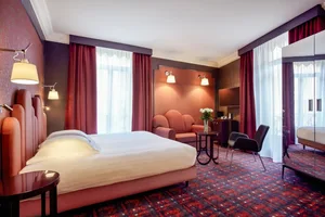 A luxurious hotel room featuring a bed, armchair, and desk with a television, all adorned with red and brown furnishings and 