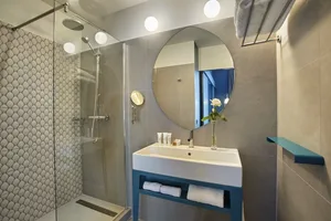 Modern bathroom with gray textured walls, concrete floor, and blue accents. Includes a circular mirror, sink, and shower.