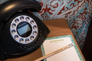 A black rotary telephone and a writing set with a pencil and paper on a wooden desk in a hotel room.