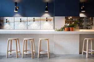 Contemporary wooden stools lined up against a sleek, white-tiled coffee shop counter with potted plants and shelves in the ba